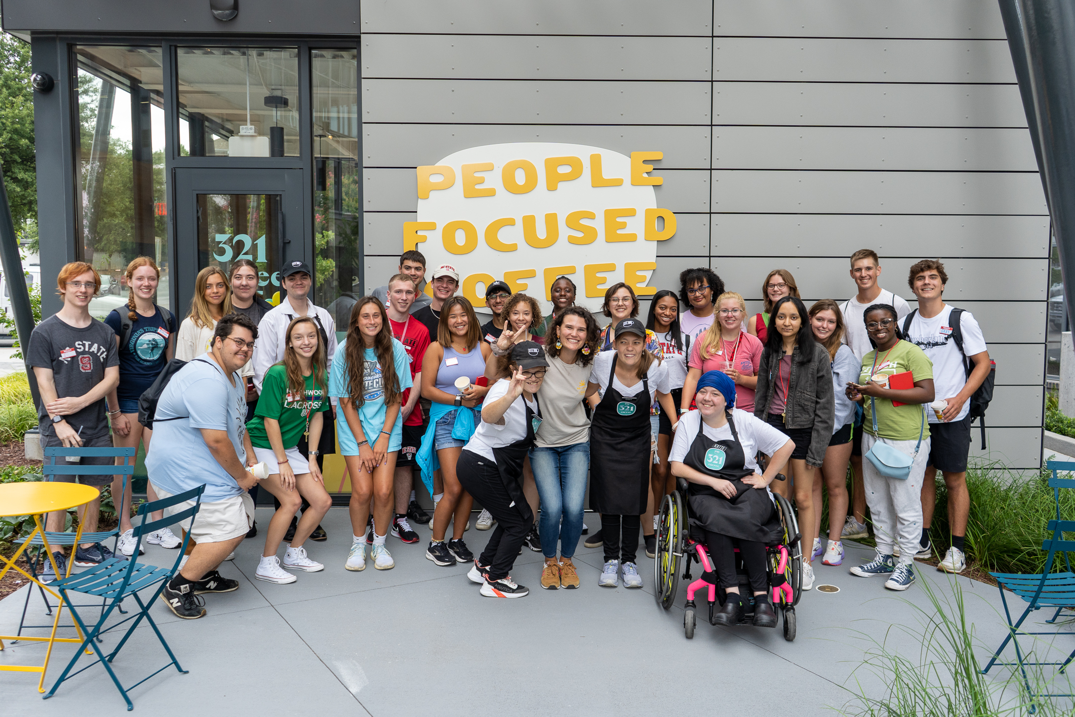 Image depicts a large group of Honors Program students in front of "People Focused Coffee," with some of that business's staff, in downtown Raleigh, NC during the Honors Program's Rooted in Raleigh summer opportunity.